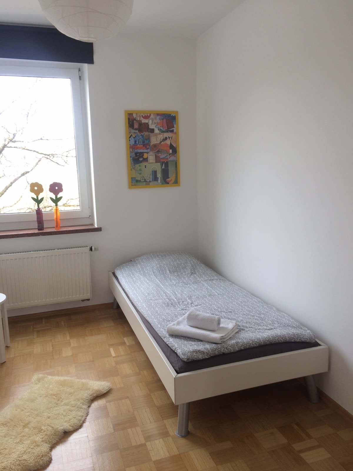 A bright room features a single bed with a gray blanket and a towel neatly folded on top. A colorful artwork is displayed on the wall above the bed, alongside two decorative figures. Natural light enters through a window dressed with a simple shade.