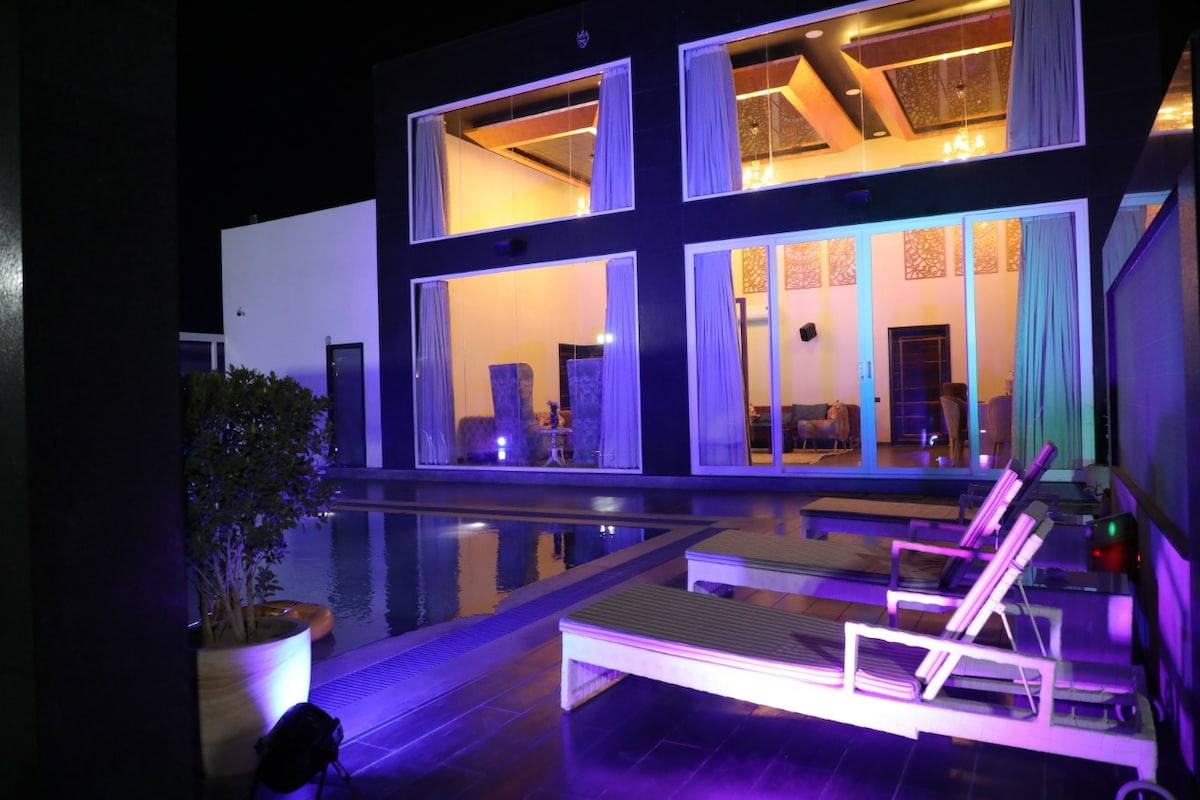 A modern villa is illuminated by soft lighting at night, showcasing large windows that reveal a spacious interior. The outdoor area features lounge chairs arranged beside a shimmering pool, reflecting the calming glow of the lights. Lush greenery enhances the tranquil setting.