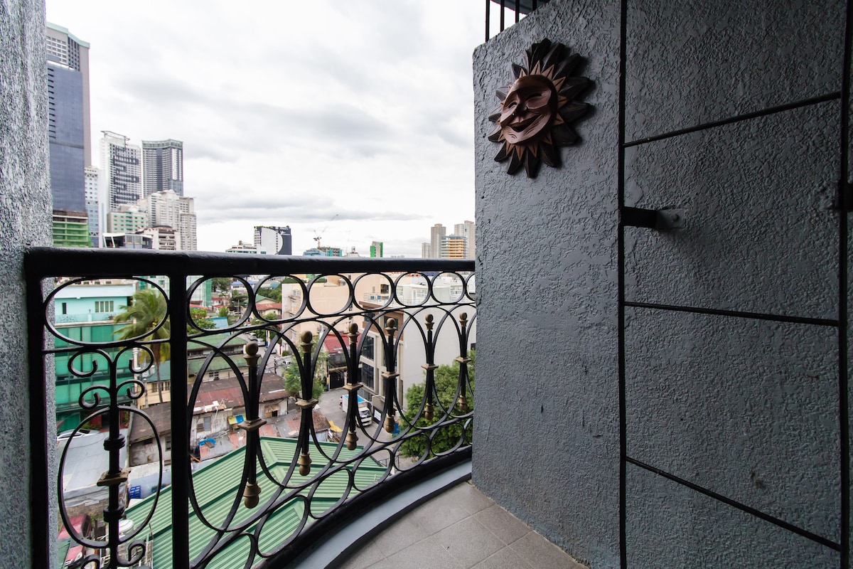 A balcony view showcases the vibrant cityscape, featuring a mix of modern buildings and greenery. Decorative wrought iron railings frame the space, while a sun-shaped wall art piece adds an artistic touch. The cloudy sky hints at a refreshing ambiance.