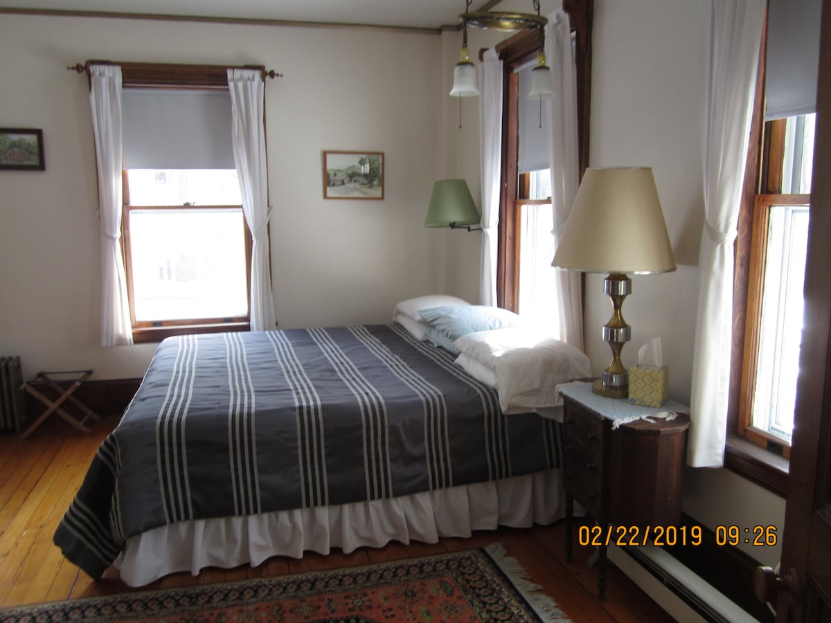 A bright bedroom features a king-sized bed with a striped blanket, flanked by two windows that allow natural light to fill the space. A wooden nightstand with a lamp and tissue box is positioned beside the bed, while a small folding chair is visible in the corner.