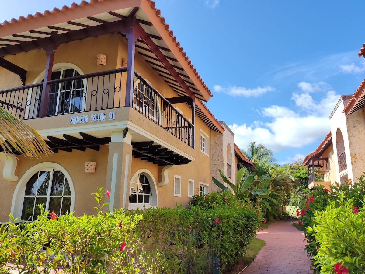 A charming apartment building is framed by lush greenery and vibrant flowers, showcasing a balcony with decorative railings. The architecture features arched windows and a warm color palette, contributing to a relaxed ambiance. Clear blue skies complement the inviting exterior.