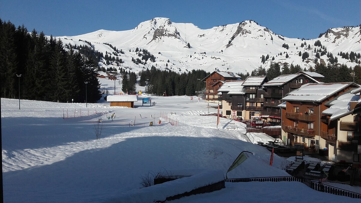 The image captures a snow-covered landscape featuring ski slopes and several chalet-style buildings. The mountains rise prominently in the background under a clear blue sky, while the foreground shows gently sloping snowfields, indicating winter activity areas.