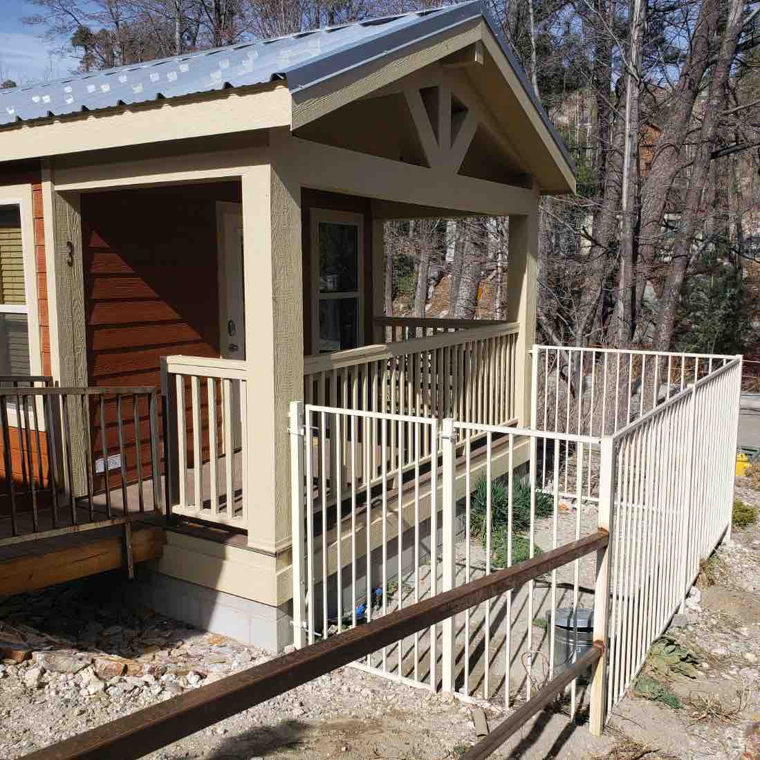 The exterior of the cabin features a covered front porch with a welcoming entrance. A railing surrounds the steps leading up to the porch, and the structure is set among trees, providing a natural backdrop.