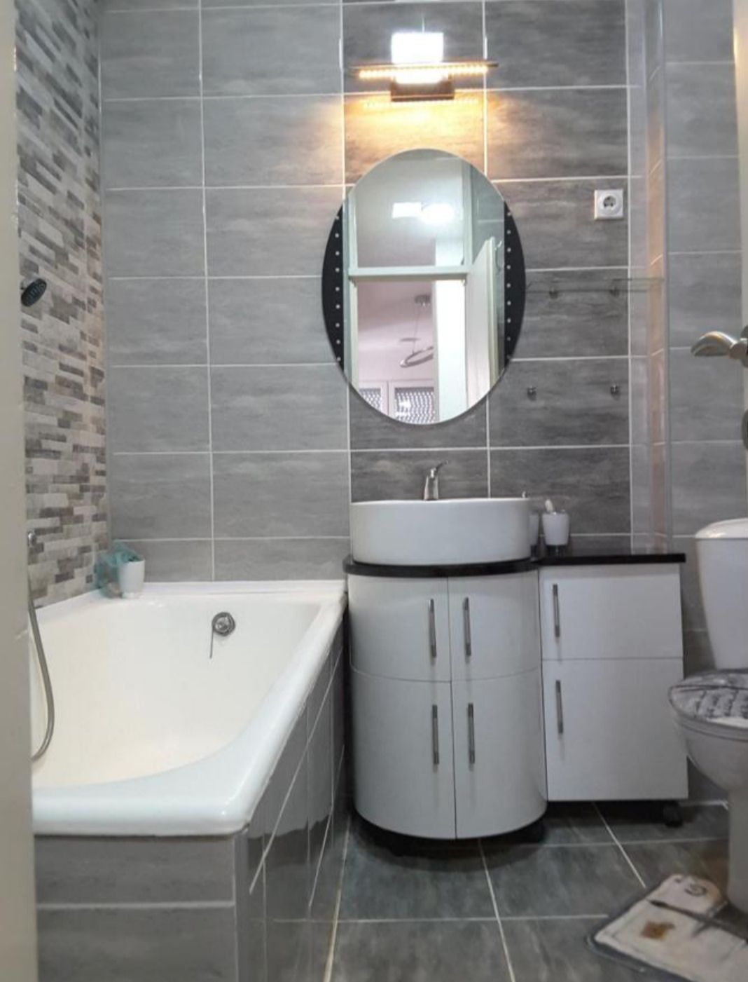 A modern bathroom features a combination of gray tile and sleek fixtures. A bathtub is positioned adjacent to a contemporary sink with under-storage. An oval mirror reflects the light above the sink, while a toilet is located in the corner.