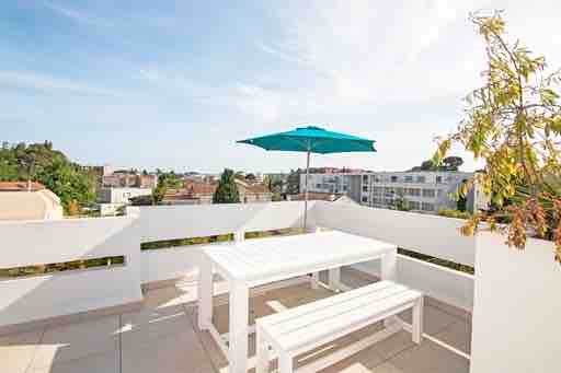 A spacious terrace features a white picnic table and benches, offering a shaded area under a bright blue umbrella. The surrounding walls provide privacy while allowing for views of the nearby neighborhood and greenery.