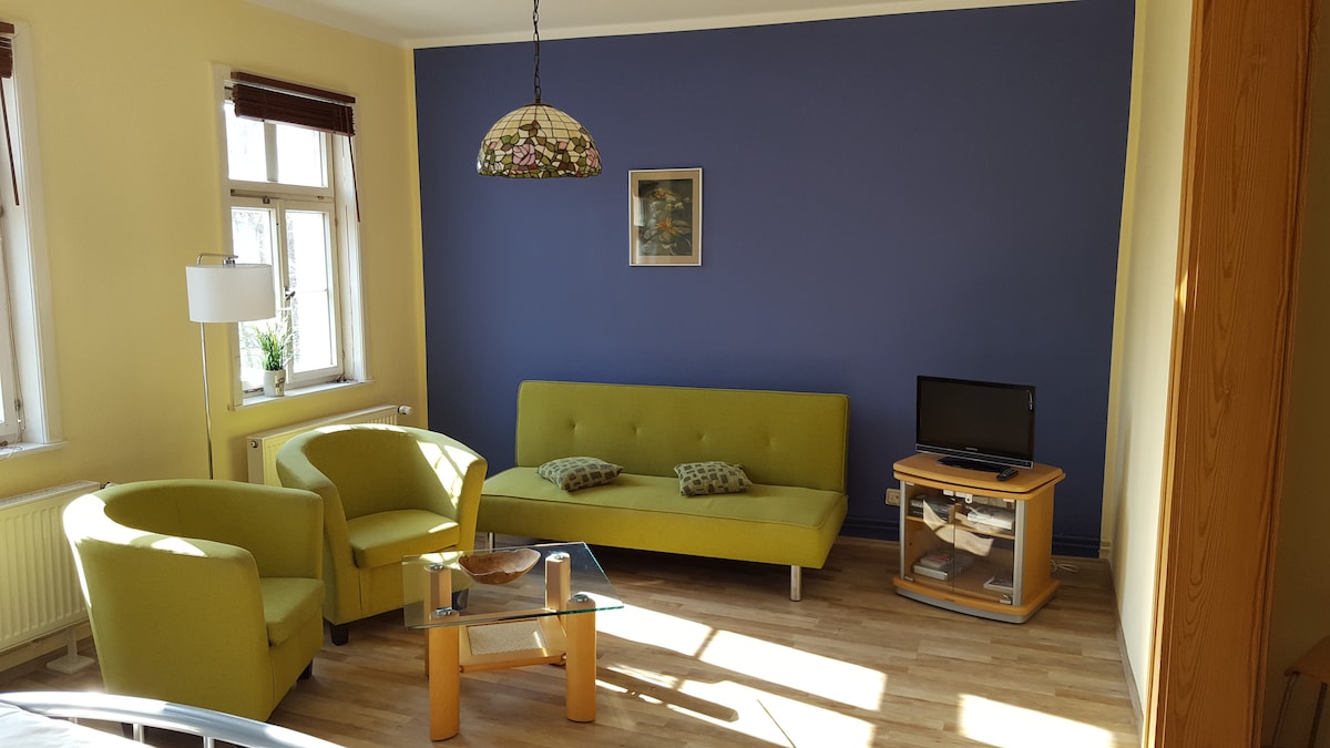 A well-lit living area features a blue accent wall and light wood flooring. Two green armchairs and a green sofa provide seating. A wooden coffee table sits in front, with a glass top and decorative items. A small television is positioned on a side table, and natural light filters through window treatments.