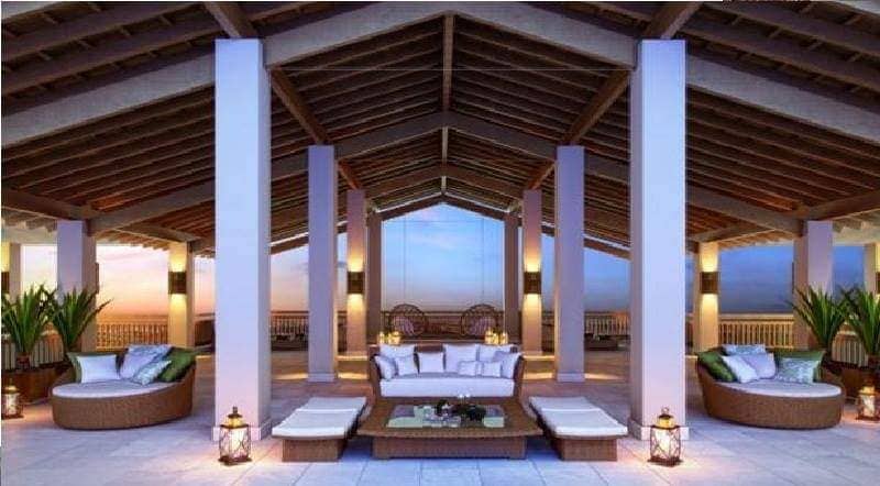 An open-air lounge area features comfortable seating arranged around a low table. Soft cushions are placed on the ground and wicker furniture provides additional seating. The space is framed by tall pillars, with plants adding greenery. Natural light fills the area, creating a calm atmosphere.
