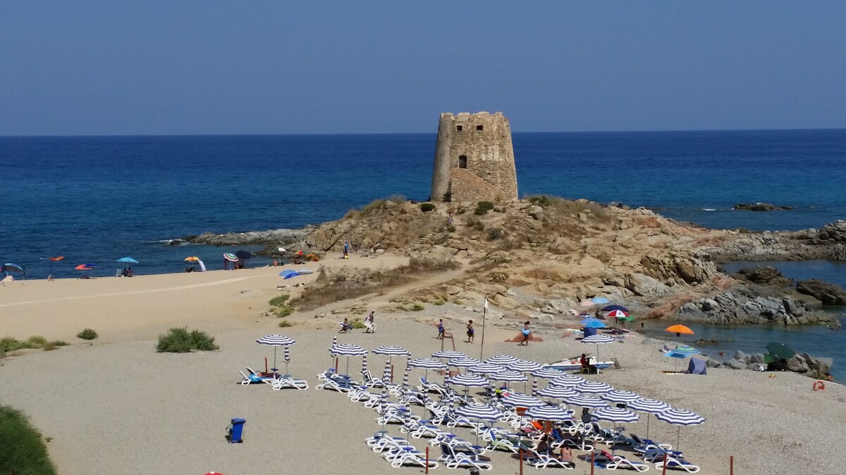 A sandy beach is visible with numerous lounge chairs arranged under striped umbrellas. A rocky outcrop features a historic tower overlooking the clear blue sea. People can be seen enjoying the beach and water, with various colorful umbrellas dotting the shoreline.