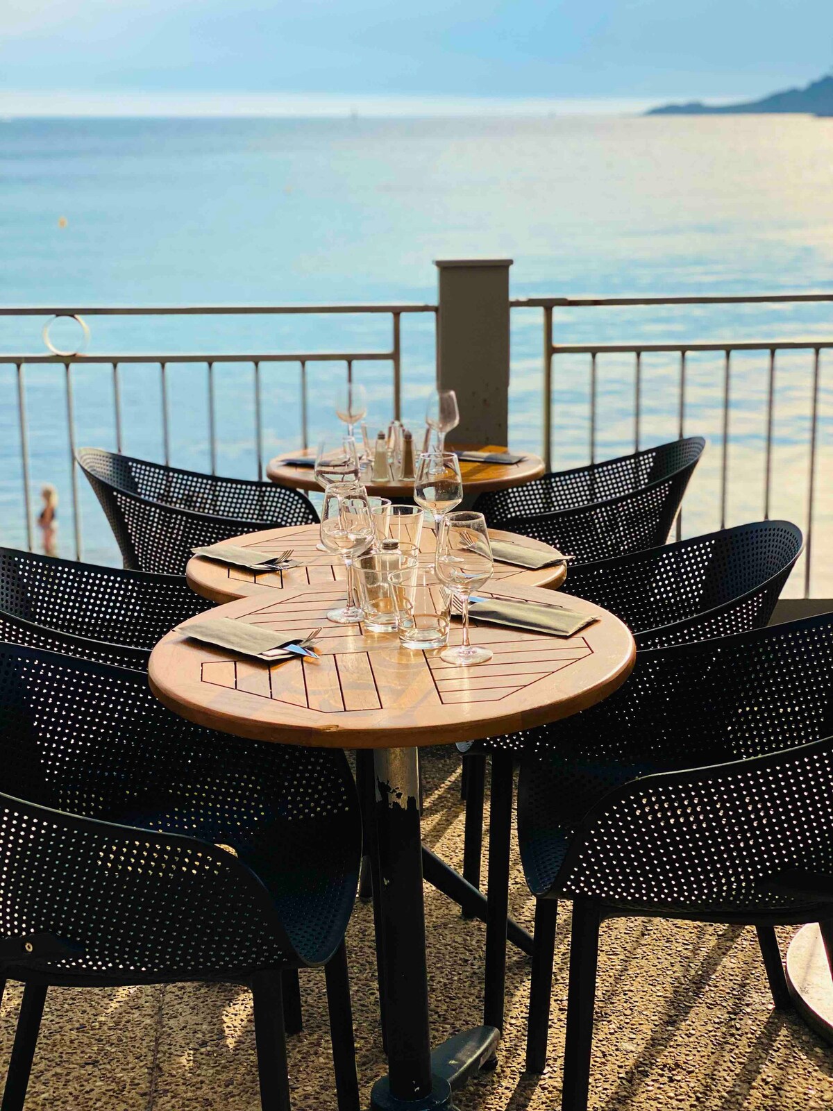 A round wooden table with a patterned design is set outside, surrounded by six black mesh chairs. Glassware is neatly arranged atop the table, with a scenic view of calm waters visible beyond the railing.