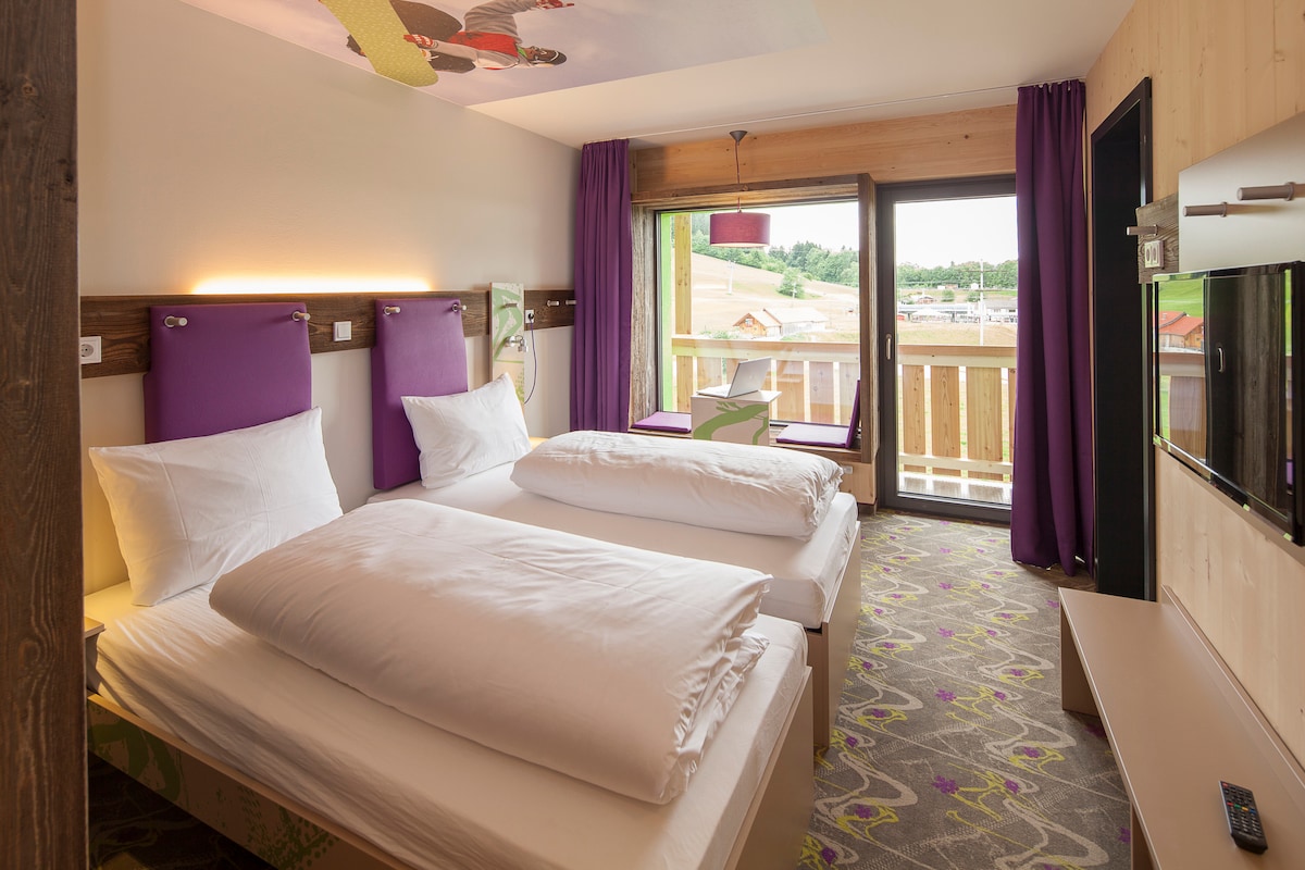 A modern double room features two spacious beds with white linens and purple accent walls. Large glass doors open to a private balcony overlooking green hills. A television is mounted on the wall, and bright natural light fills the room, complemented by purple curtains.