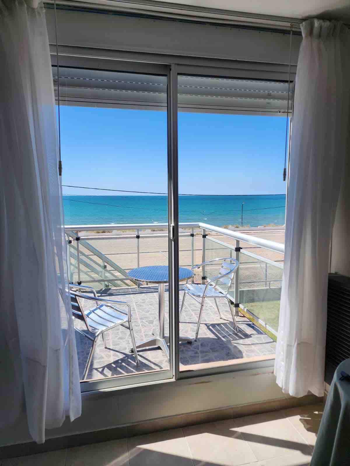 A balcony view is presented through large glass doors, showcasing the ocean and sandy beach beyond. Two chairs and a small table offer an inviting space for relaxation. Bright blue skies reflect a serene atmosphere, with natural light illuminating the interior.
