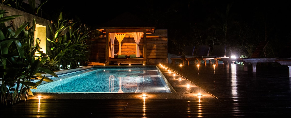 An inviting outdoor area features a well-lit swimming pool, bordered by softly glowing lights. A wooden pavilion with draped fabric sits in the background, surrounded by lush greenery, creating a serene environment for relaxation.