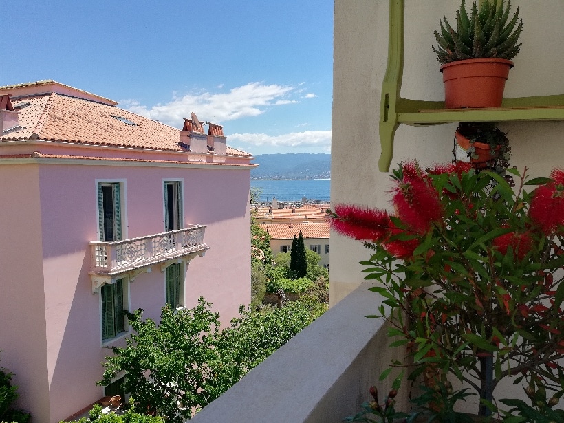 A view from the balcony showcases a vibrant pink building and the surrounding greenery. A potted plant adds a touch of nature, while the distant sea and mountains create a pleasant backdrop under a clear blue sky.