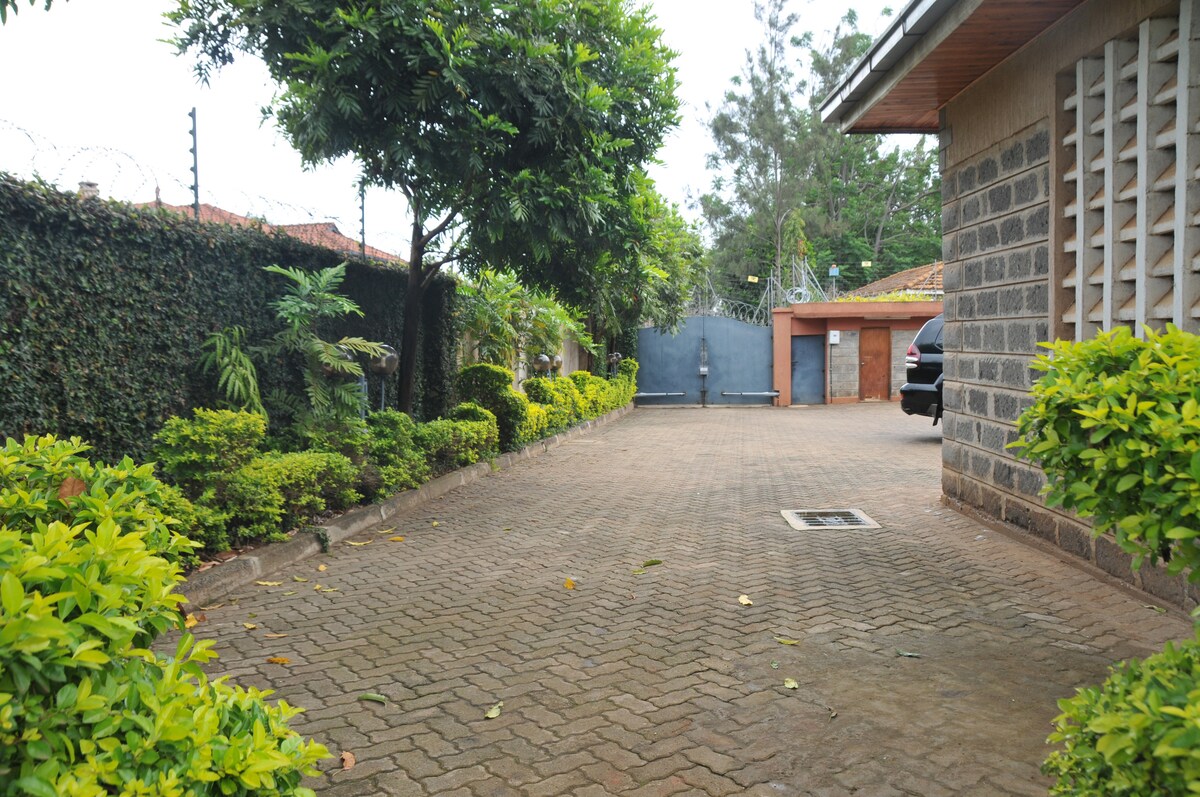 A private driveway is flanked by well-maintained greenery and low hedges. The pathway is paved and leads to a secure entrance. In the background, a large gate is visible, providing access to the property, situated in a peaceful outdoor setting.