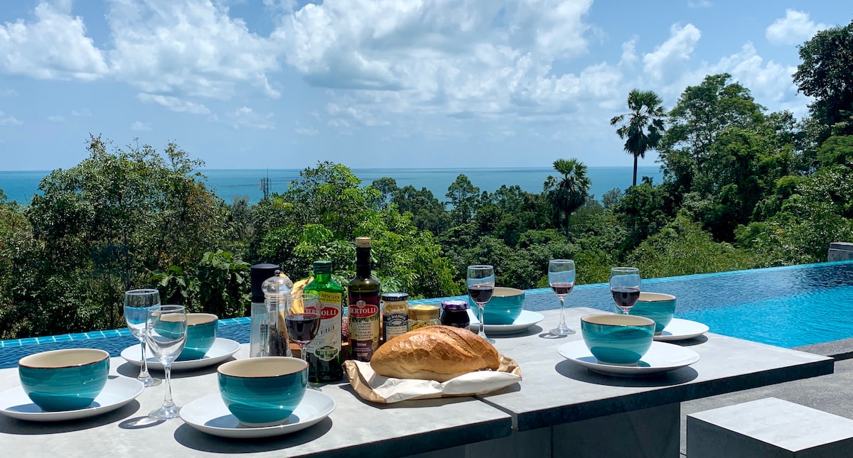A dining setup is presented by a glistening infinity pool, with six turquoise bowls arranged on a stone table. A freshly baked loaf and various condiments accompany glasses filled with beverages, all framed by lush greenery and distant ocean views under a bright sky.