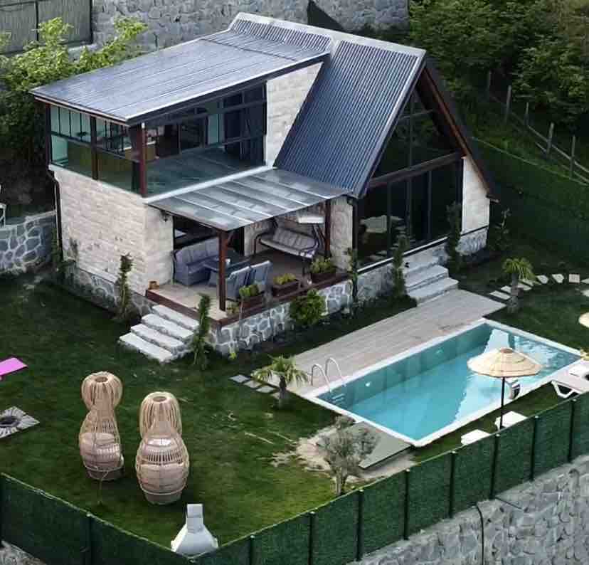 Aerial view of the property reveals a modern house with a large glass facade, surrounded by a well-kept garden. The outdoor pool is visible alongside lounge chairs, while two woven chairs add a cozy touch to the space.