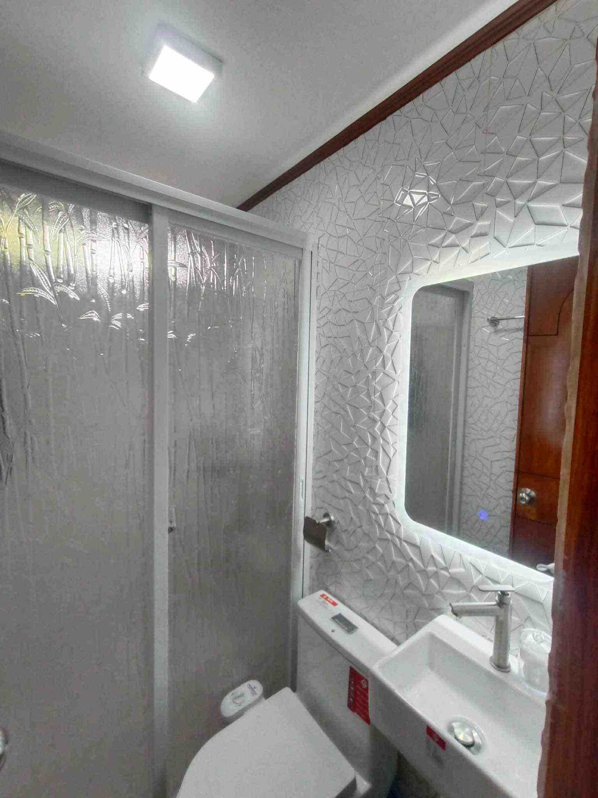 A modern bathroom is presented, featuring a glass shower enclosure and a mirrored vanity with integrated lighting. The textured wall adds dimension, while the toilet and sink are positioned for accessibility. Bright overhead lighting ensures a well-lit space, enhancing usability.
