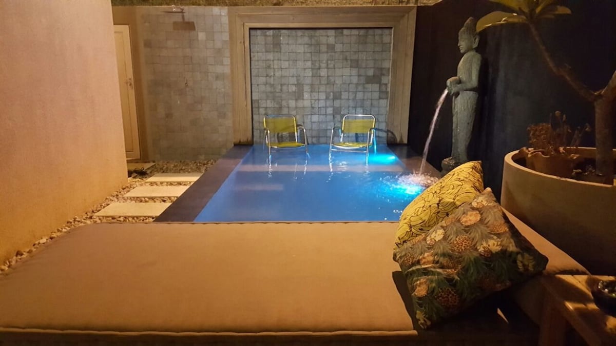 A serene pool area is depicted, featuring a crystal-clear private pool illuminated with soft blue lighting. Two modern chairs are positioned at the pool's edge, while a soothing water feature adds tranquility. Decorative plants and artwork enhance the space's ambiance.
