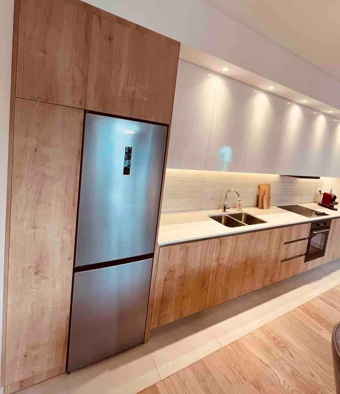 A modern kitchen features sleek cabinetry in light wood and white finishes. Stainless steel appliances, including a refrigerator and oven, are neatly integrated. Counter space includes a double sink along with wooden cutting boards, providing a functional and inviting area for meal preparation.