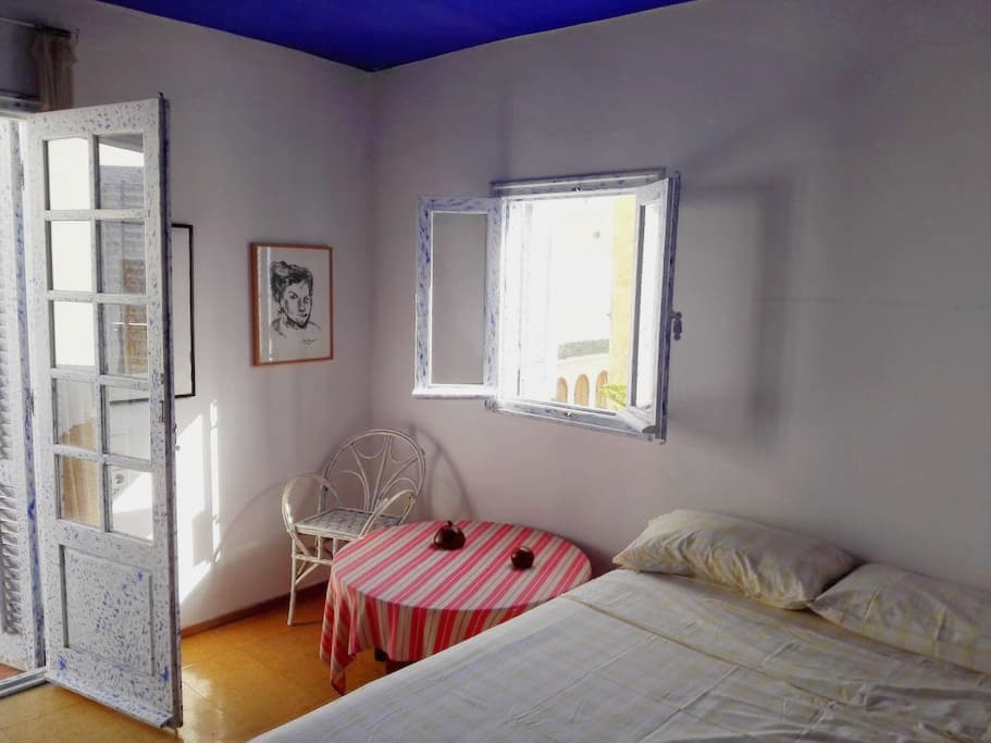 A bright room features a spacious bed and a small round table with striped cloth. A white, wicker chair complements the setup near a large, open window. Sunlight streams in, illuminating the fresh white walls and vibrant blue ceiling.