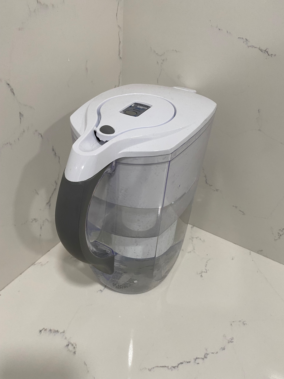 A water pitcher with a sleek design is situated against a marble surface. It features a white lid and a gray handle, showcasing its modern aesthetic. The transparent body reveals the water level inside, providing easy visibility.