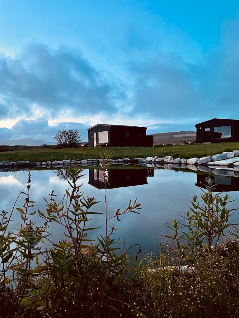 Two modern lodges are positioned beside a tranquil pond, reflecting their dark wood exteriors under a dusky sky. Lush greenery and rocks frame the pond, enhancing the serene landscape surrounded by distant hills.