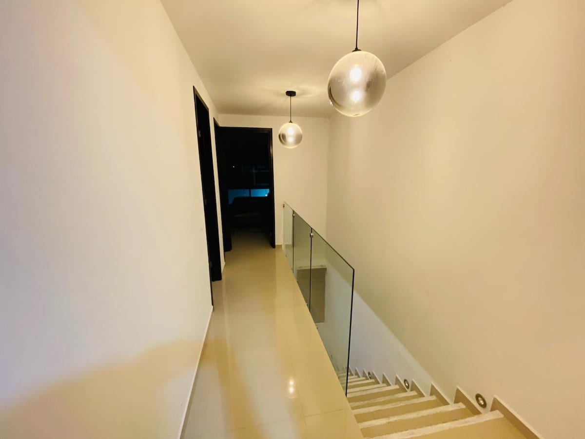 A well-lit hallway is shown, featuring sleek white walls and a glossy tiled floor. Two modern pendant lights hang from the ceiling, providing illumination. A clear glass railing borders the staircase, leading down toward a darker space in the background.