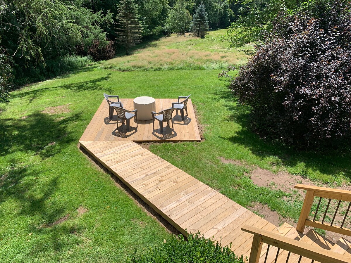 A wooden deck area is visible, featuring four chairs arranged around a circular table. The space is surrounded by lush green grass and trees, creating a peaceful outdoor setting. A wooden pathway leads to the deck, enhancing accessibility.