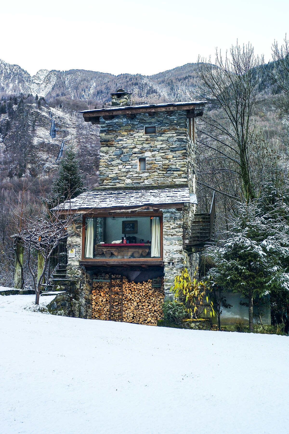 A historic stone tower, partially covered in snow, is surrounded by trees and mountain views. A large glass window on the lower floor showcases a cozy interior. Stacked firewood is neatly arranged at the entrance, adding a rustic touch to the inviting facade.