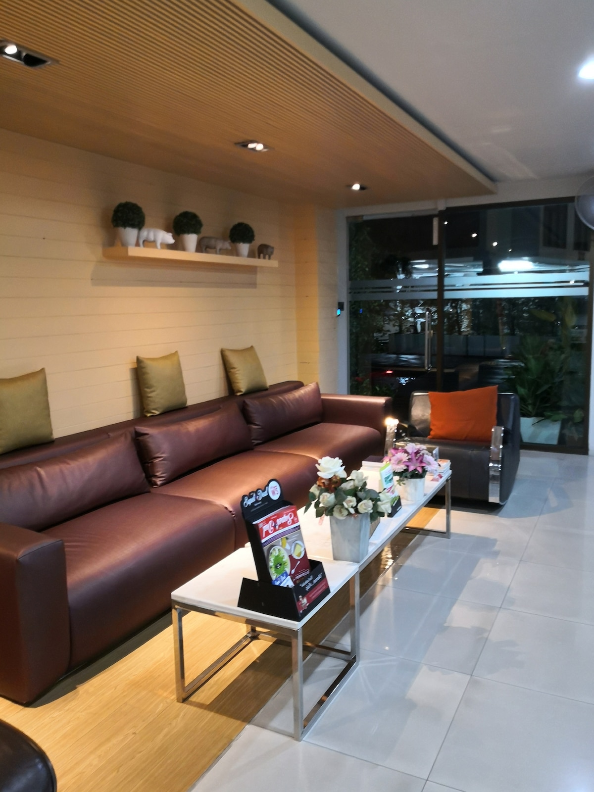A modern living area is presented with a brown leather sofa and silver accent chairs. A glass coffee table is centered between them, adorned with a small flower arrangement. Natural light filters in through large windows, creating an inviting atmosphere.