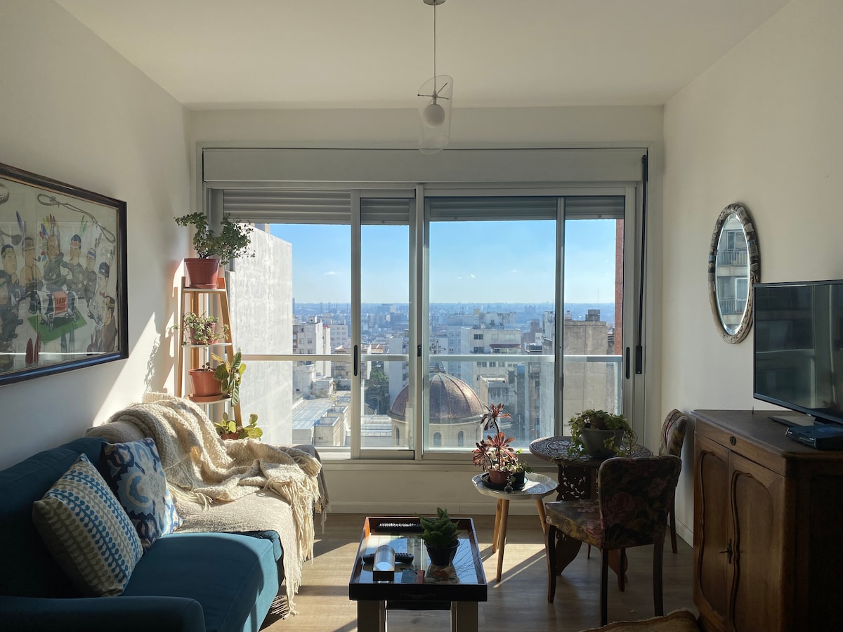 A bright living area is highlighted by large windows that reveal expansive city views. A cozy blue couch is complemented by decorative pillows and a soft throw. A wooden coffee table is placed between two chairs, with plants enhancing the space's natural feel.
