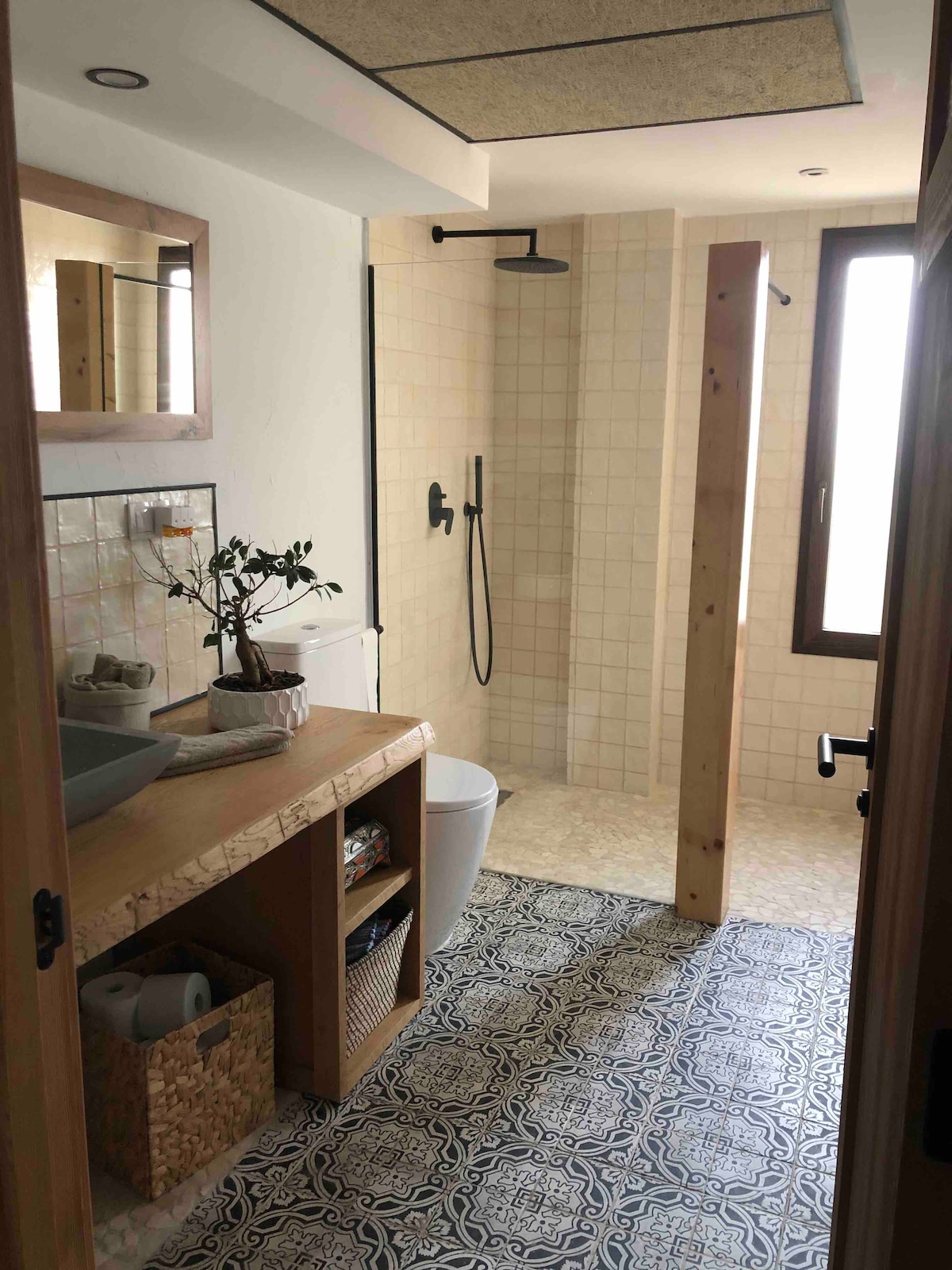 A modern bathroom is presented with natural wood accents. A freestanding sink is located beside a woven basket. The spacious shower area features a rainfall showerhead and tiled walls. Light enters through a window adjacent to the shower, adding brightness to the room.