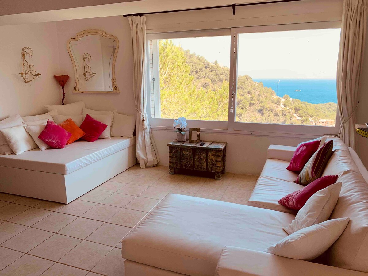 A bright living area is furnished with a large white sectional sofa and a comfortable daybed, accented by colorful cushions in shades of red and pink. Large windows frame stunning views of the sea and surrounding hills, enhancing the tranquil ambiance.