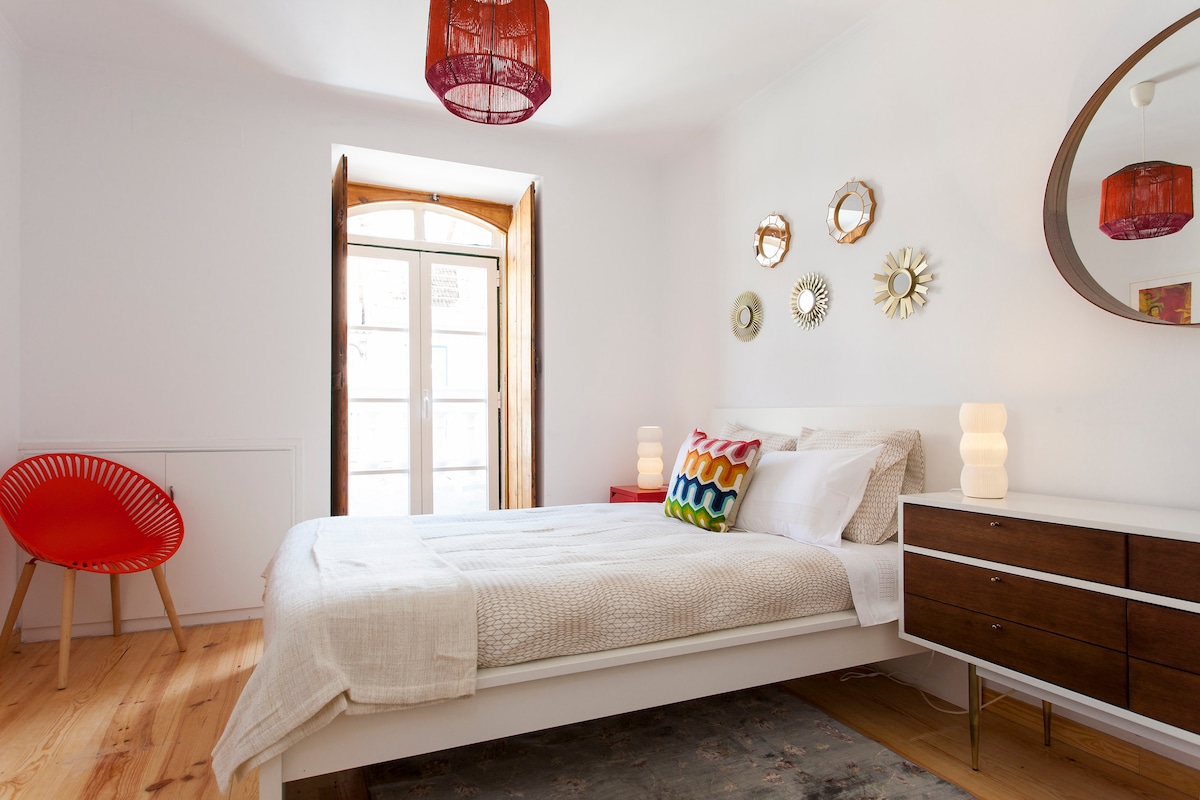 A bright bedroom features a spacious bed with soft bedding, complemented by colorful decorative pillows. Large windows with wooden frames provide natural light, while a modern chair adds a pop of color. Stylish mirrors adorn the wall, enhancing the room's inviting atmosphere.