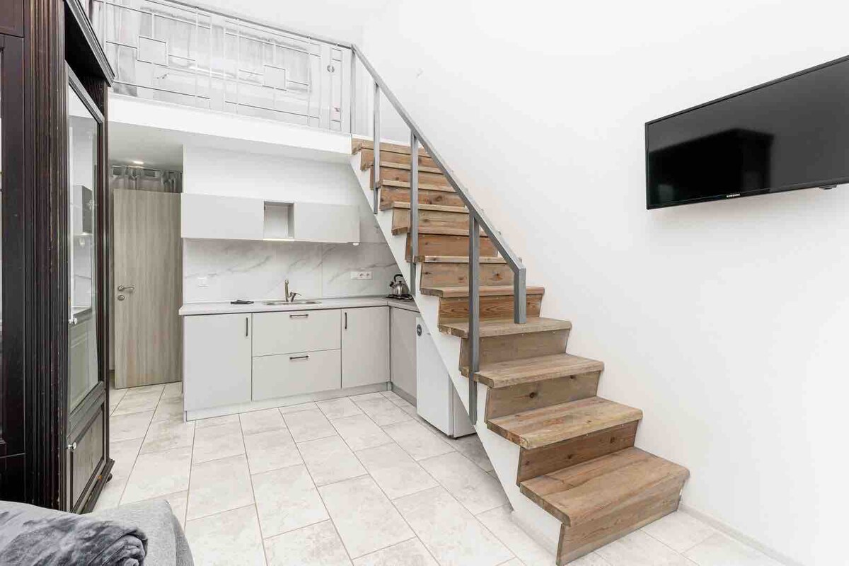 A modern interior features a staircase with wooden steps leading to an upper level. A compact kitchen area is visible, equipped with sleek cabinetry and minimalistic design elements. Light-colored tiles cover the floor, enhancing the space's openness, while a wall-mounted television is positioned nearby.