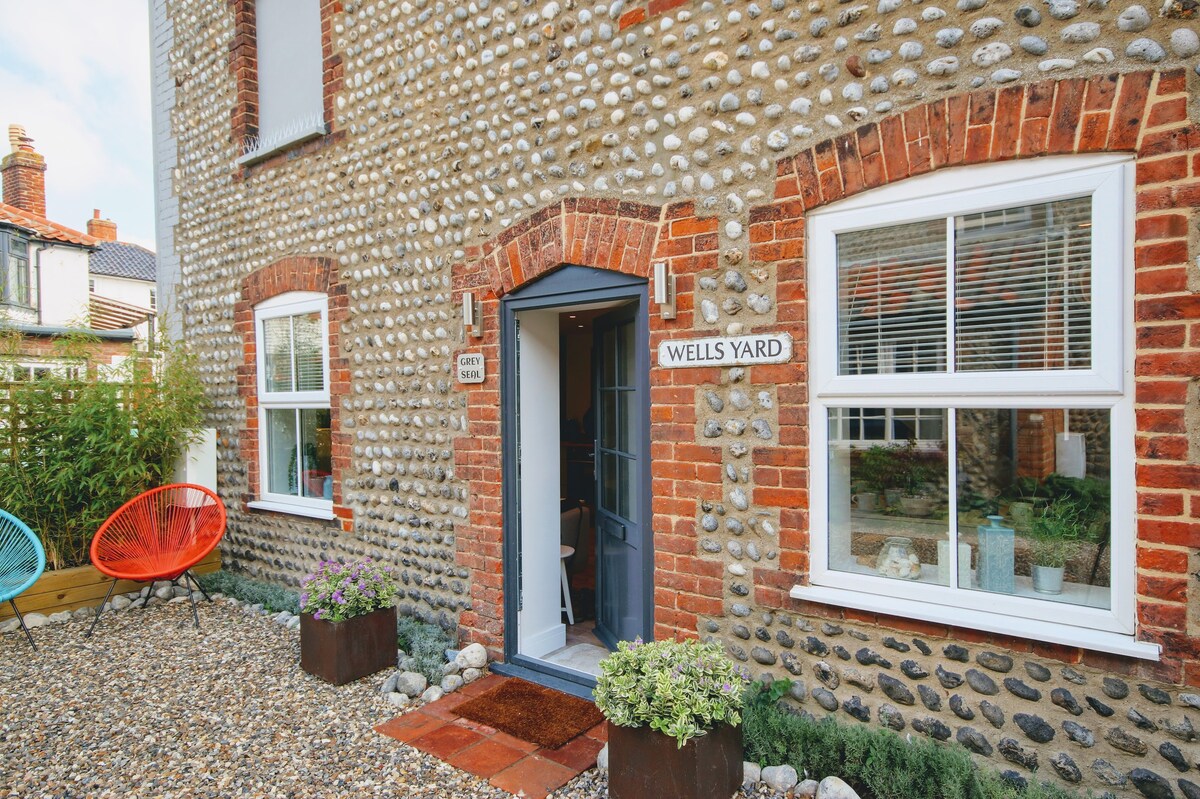 The entrance of the townhouse features a charming blue door framed by smooth, round pebbles and red brick. A small garden area is visible, adorned with colorful chairs and planters filled with greenery. The nameplate 'Wells Yard' is mounted beside the front door.