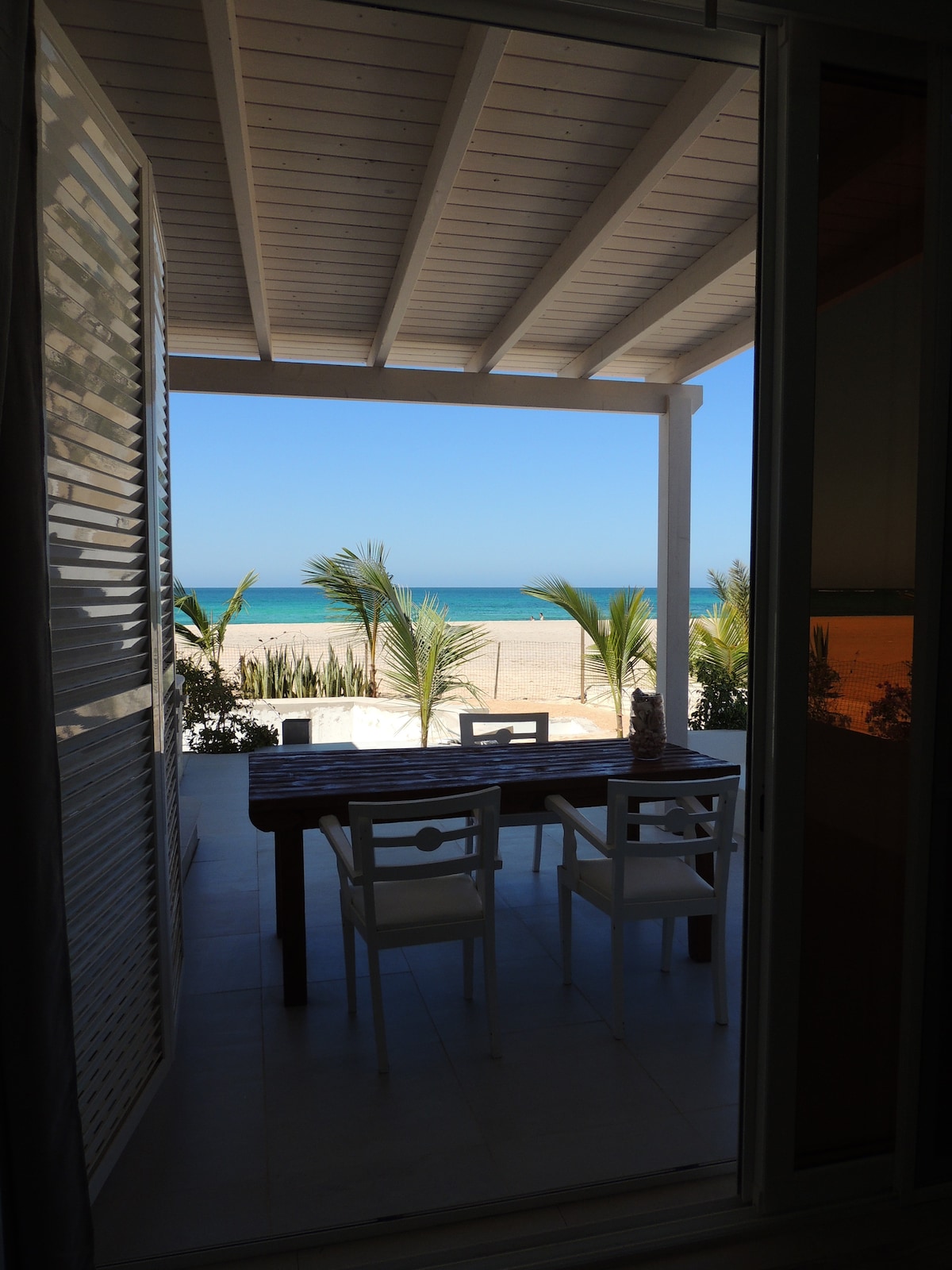 A private veranda is shown with a table and chairs, framed by large glass doors. Beyond the veranda, the sandy beach and turquoise ocean are visible, accented by palm trees lining the shore under a clear blue sky.