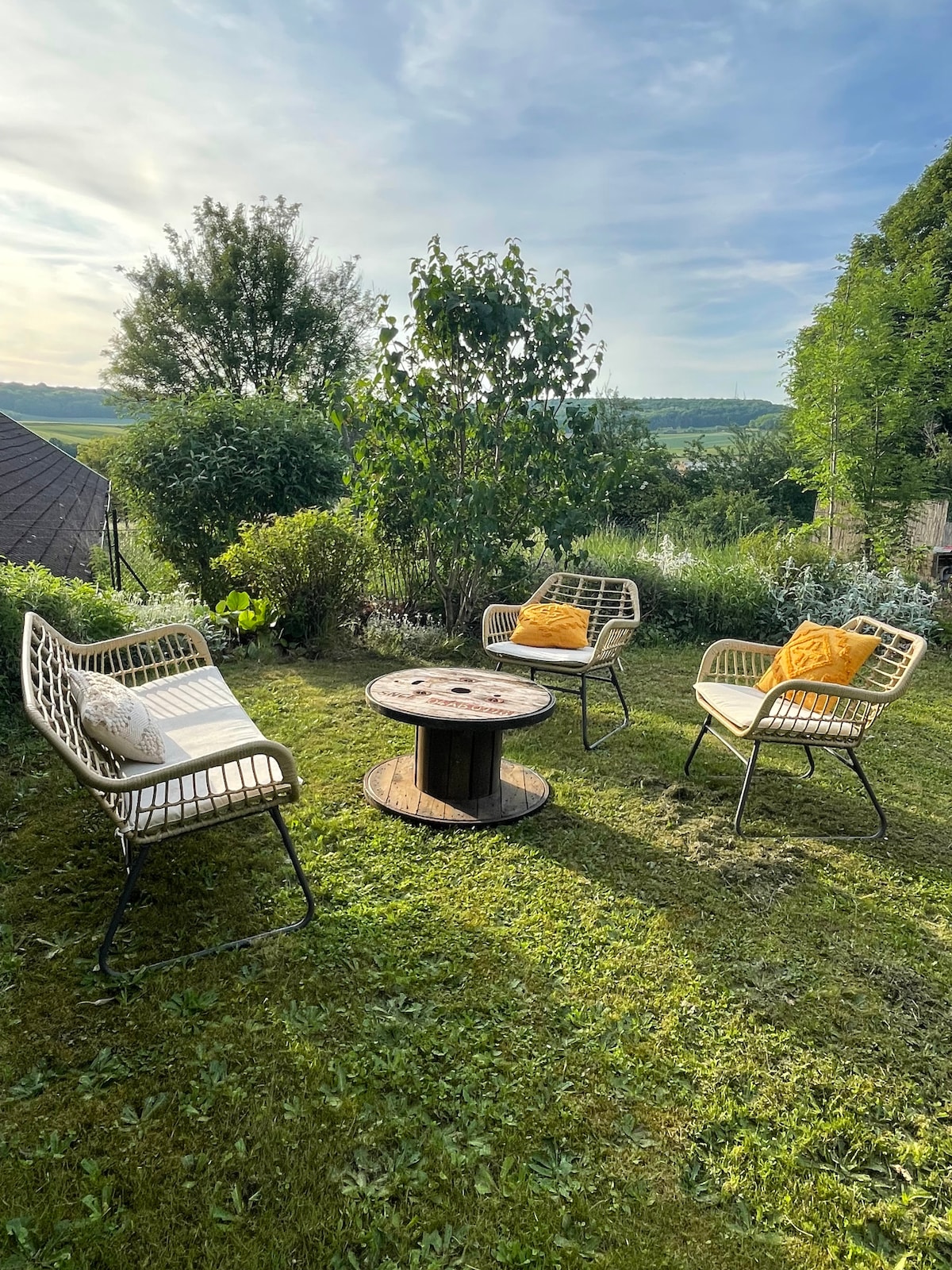 A serene outdoor space features a circular table surrounded by four comfortable chairs. Soft cushions add a pop of color, while lush greenery and distant hills create a peaceful backdrop. The atmosphere invites relaxation under the open sky.