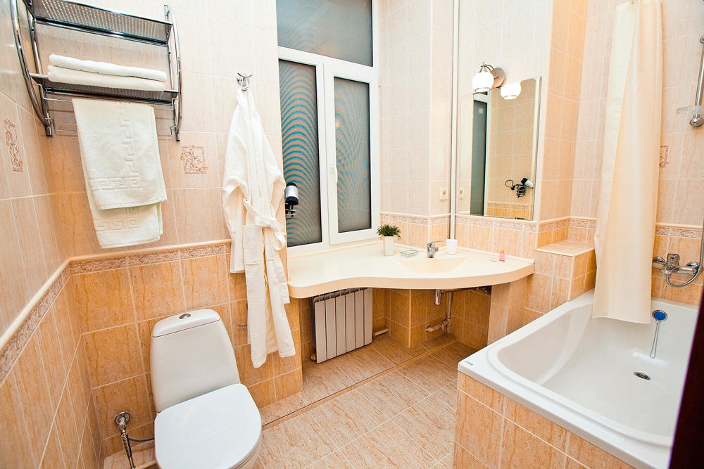 A well-appointed bathroom features beige tiled walls and flooring. A spacious sink counter runs beneath a large window. A curved bathtub is positioned next to the toilet. Fresh towels are neatly arranged on a shelf, complementing the overall clean and functional design.
