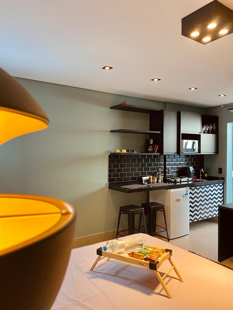 A compact kitchen area is detailed with black cabinetry and contrasting countertops. An electric two-burner stove, microwave, and mini fridge are visible. Minimalist bar stools accompany the counter. Soft lighting illuminates the space, enhancing a functional and well-organized cooking environment.
