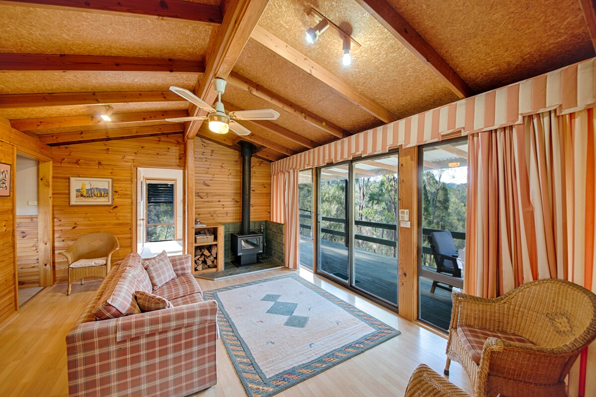 A spacious living area features wooden beams and large windows that overlook the surrounding bushland. A comfortable sofa and armchairs are arranged around a central rug. A fireplace adds a cozy touch, while sliding doors provide access to the outdoor deck.