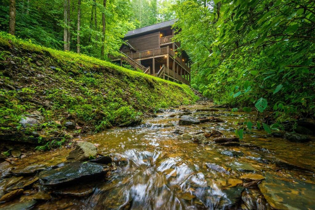 The cabin is set among lush greenery, surrounded by trees and located beside a gently flowing creek. Smooth rocks create a natural pathway along the water's edge, complementing the serene outdoor environment. The cabin's entrance is visible above, blending harmoniously with its wooded surroundings.