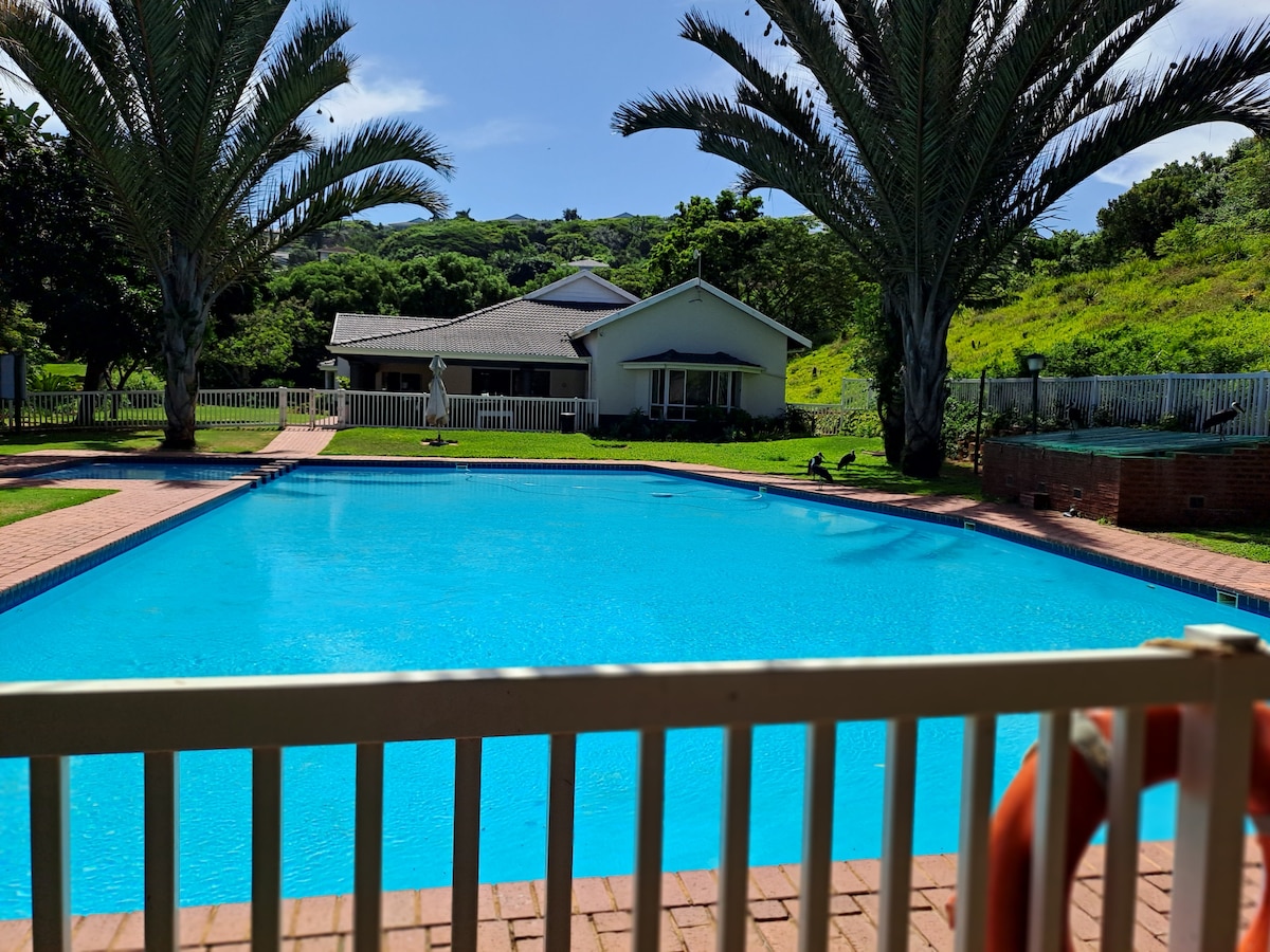 A large pool is centered within the estate, surrounded by a deck and lined with tall palm trees. A spacious house is visible in the background, framed by lush green hills. The tranquil water reflects the bright blue sky.