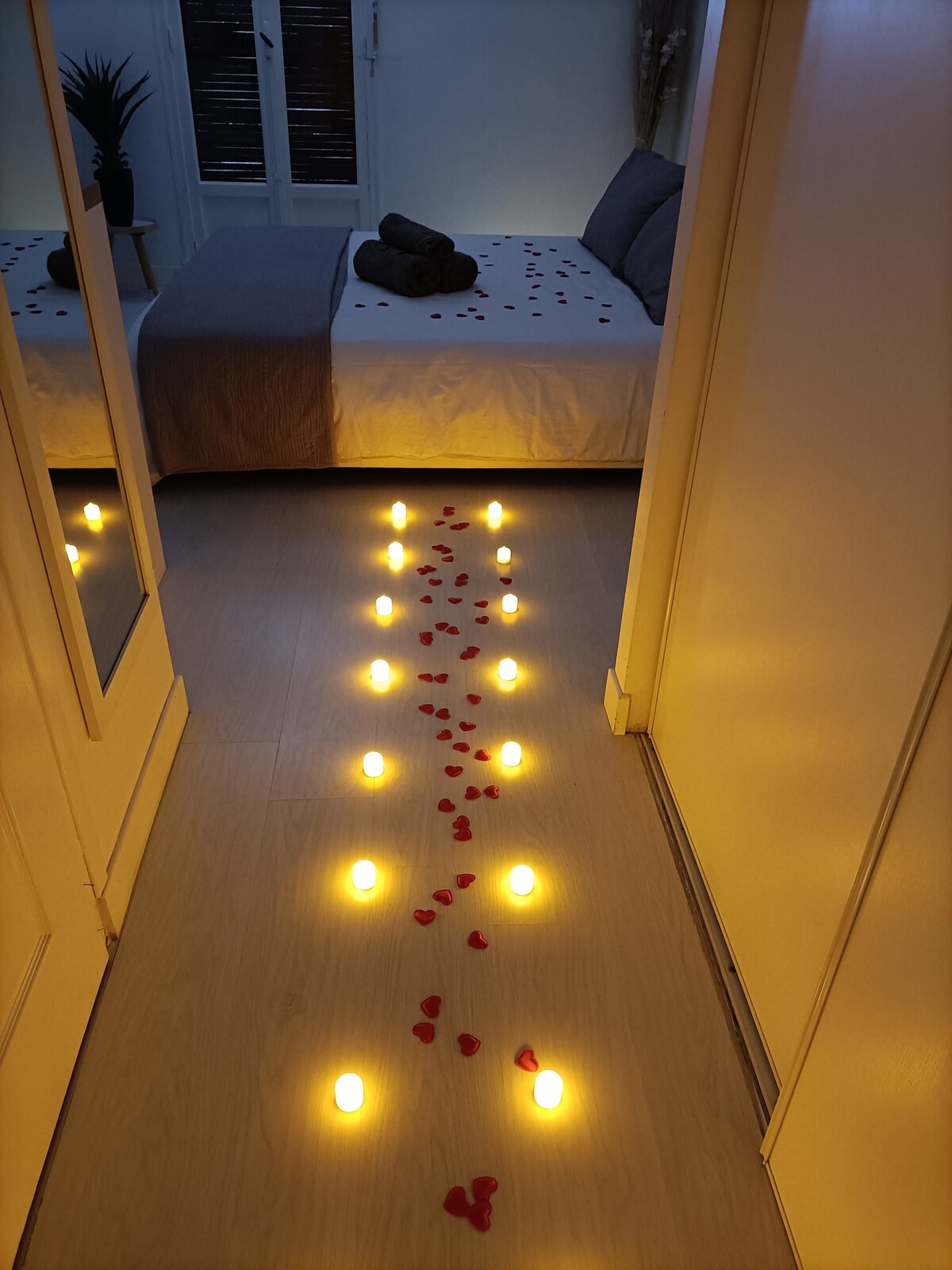 A serene indoor pathway is created with illuminated candles and rose petals, leading to a beautifully arranged bed. Soft lighting highlights a cozy environment, while a towel set and decorative touches are visible on the bed, inviting relaxation.