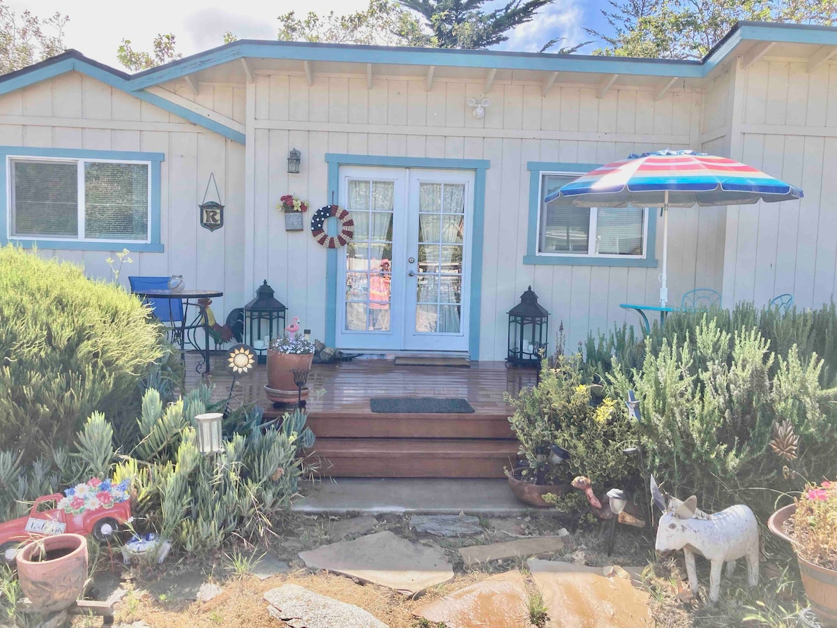 The entrance of a charming cottage is framed by lush greenery and blooming flowers. Blue and white accents are present on the door and furniture. A colorful umbrella provides shade over a small outdoor seating area, while decorative lanterns and garden art add character to the scene.