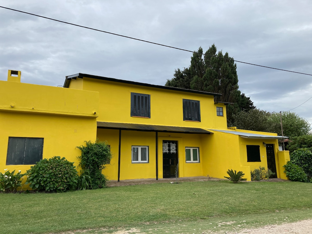 The exterior of a two-story yellow house is visible, surrounded by greenery. Large windows offer natural light, while the entrance features a central door flanked by smaller windows. The well-maintained lawn adds to the inviting atmosphere of the property.