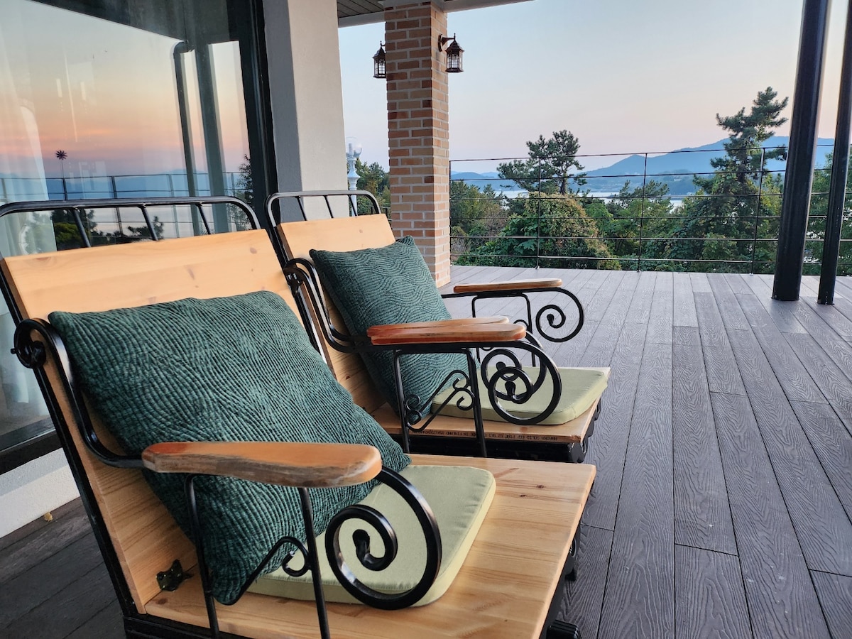 Two wooden lounge chairs with dark green cushions are positioned on a spacious terrace. A panoramic view of the sea and surrounding nature can be seen in the background, complemented by soft, warm hues from the setting sun.