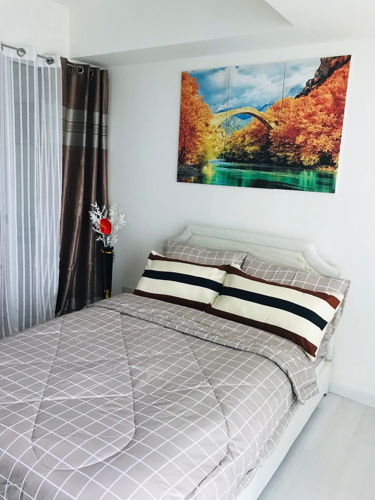 A serene bedroom is seen featuring a spacious bed adorned with patterned bedding and decorative pillows. A large artwork depicting an autumn landscape hangs above the bed. Sheer curtains frame the window, allowing natural light to filter in softly.