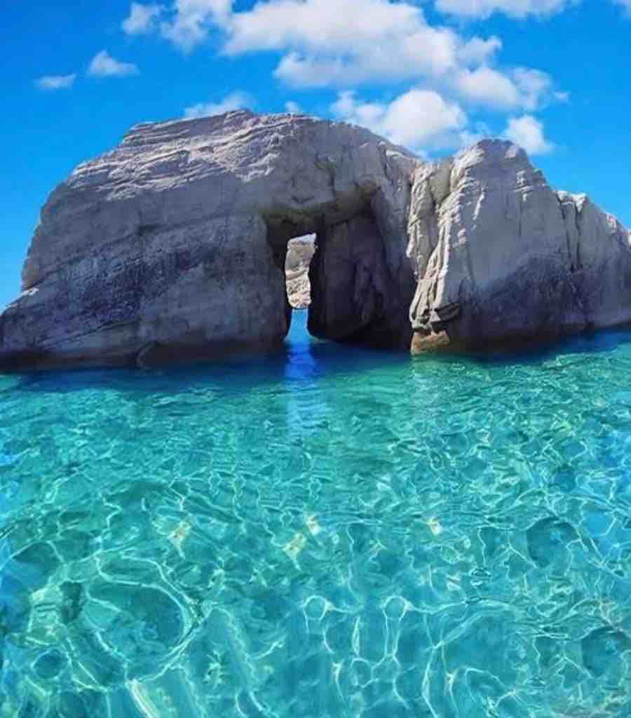 A natural rock formation is seen above the crystal-clear turquoise waters. The opening in the stone is framed by smooth, weathered cliffs, reflecting the vibrant blue sky dotted with fluffy, white clouds. The shimmering water reveals a beautiful underwater texture.