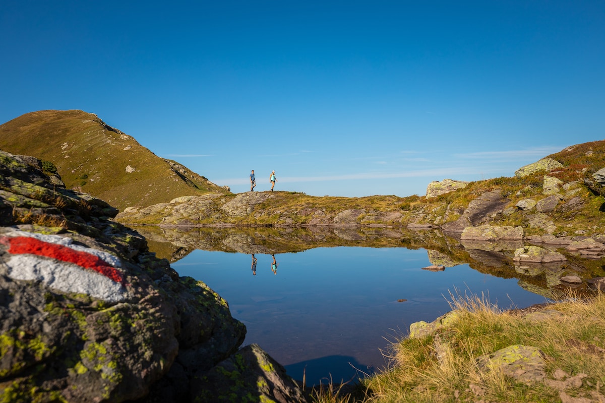 Two individuals are seen walking along a scenic path by a calm pond, reflecting the vibrant blue sky above. Surrounding hills are gently rolling, with patches of greenery and rocks visible in the foreground, creating a serene natural landscape.
